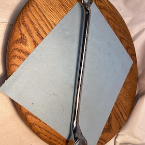 Op de afbeelding: Een verchroomde steeksleutel, met een ringkant en een open kant, ligt op een lichtblauwe papieren handdoek. De sleutel ligt diagonaal over een ronde houten ondergrond met zichtbare houtnerf.