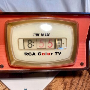 May include: A vintage RCA Color TV flip clock with a coral-coloured plastic frame and a cream-coloured face. The display shows the text TIME TO SEE... above the numbers 8:55 and a small vertical dial with increments of 5, 10, and 15.