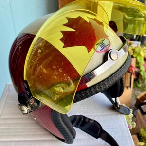 May include: A motorcycle helmet featuring a cream and maroon exterior with a prominent red maple leaf graphic. The helmet includes a translucent yellow flip-up visor attached with silver metal hardware and a black chin strap with padding.