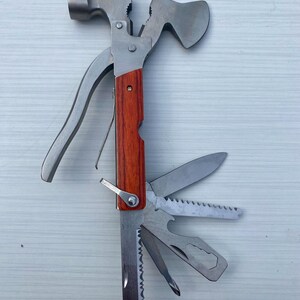 May include: A multi-tool featuring a silver hammer head and hatchet blade at the top, with a wooden handle grip. The base includes multiple fold-out stainless steel implements, including a knife blade, a saw, a bottle opener, and a screwdriver.
