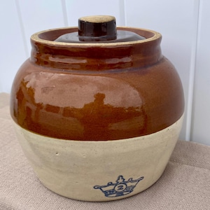May include: A round stoneware crock with a lid, featuring a two-tone design with a glossy brown upper section and a matte cream lower section. A blue stamped crown logo with the number 2 inside is visible on the lower front of the vessel.