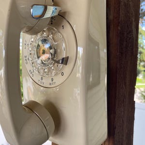 May include: A cream-coloured wall-mounted rotary telephone featuring a circular dial with numbers zero through nine and corresponding letters. The handset rests on a chrome-finished cradle hook, and a coiled cord extends from the bottom of the unit.