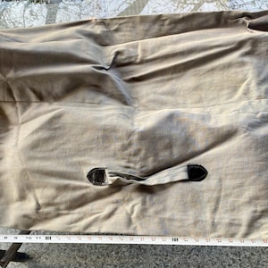 May include: A rectangular, light brown canvas storage bag featuring a central fabric handle with black leather-like accents and a drawstring closure with metal grommets on the right side. A white measuring tape shows a length of 71 cm.