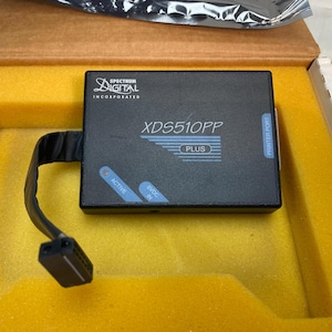 May include: A black XDS510PP PLUS device with a cable and connector, inside a yellow foam-lined box. The device has the Spectrum Digital Incorporated logo and the words "ACTIVE" and "5VDC IN" in blue.