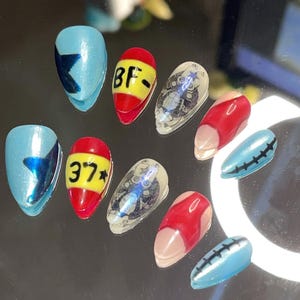 May include: Set of ten false nails with various designs.  Some nails are blue with a star design, some are red with yellow numbers and a star, some are clear with a black and white design, and some are red with a red design.