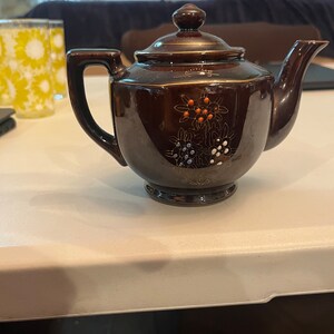 May include: A brown teapot with a floral design in gold, white, and red. The teapot has a lid and a spout.