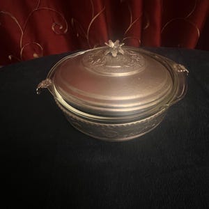 May include: A silver-colored casserole dish with a glass base and a decorative lid. The lid features a floral design and a flower-shaped handle. The dish has handles on either side and is set against a dark background.