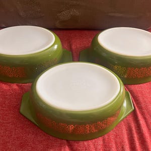 May include: Three vintage Pyrex bowls in a vibrant green hue, each featuring a white interior and a decorative orange floral pattern. The bowls have a rounded shape with handles on either side, perfect for serving or storage.