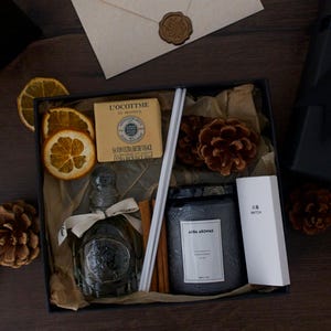 Op de afbeelding: Een zwarte cadeaudoos gevuld met verschillende items, waaronder een fles parfum, een kaars, een zeep en een set diffuserstokjes. De doos is omringd door dennenappels en gedroogde sinaasappelschijfjes.