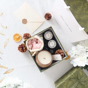 May include: A green gift box with a lid that says "Just for you" contains a pink crystal, a coconut shell candle, and two small glass bottles with black lids. The box is sitting on a white surface with a white envelope and a small orange.