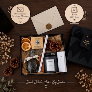 Op de afbeelding: Een geschenkdoos met een stuk L'Occitane-zeep, een glazen fles met een wit lint, twee gedroogde sinaasappelschijfjes, dennenappels, kaneelstokjes, een zwarte Aura Aromas-kaars en een wit doosje lucifers, inclusief een envelop met een zegel.
