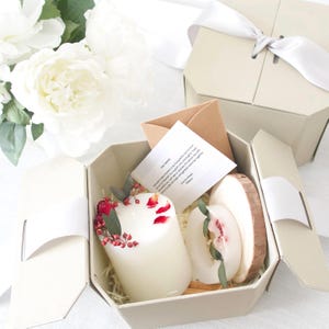May include: A white gift box with a white ribbon and bow. The box is open and contains a white candle with red flowers and a wooden slice with a white candle on top. The box also contains a small card with a handwritten message.