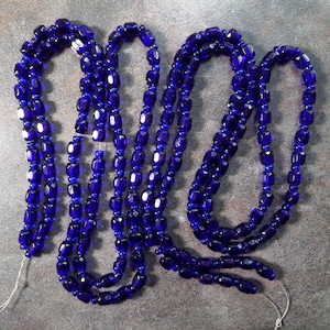 May include: Several strands of deep blue glass beads. The beads are oblong and faceted, strung together with a thin, white string. The beads have a glossy finish and reflect light.