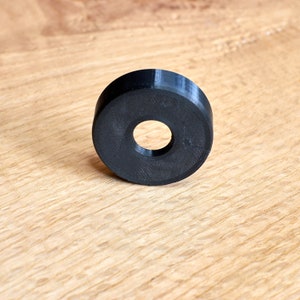 May include: A black plastic ring with a hole in the center. The ring is lying on a light brown wooden surface.