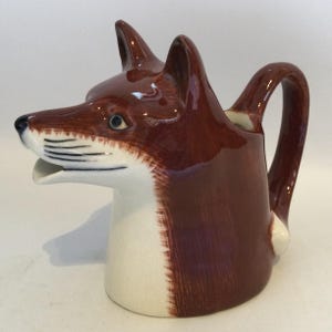 May include: A ceramic creamer jug shaped like a fox head. The jug features a glossy brown and white finish, with painted black whiskers and a black nose. The spout is integrated into the fox's mouth, and a curved handle extends from the back.