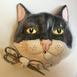 Puede incluir: Una maceta de pared de cerámica con forma de cabeza de gato con un patrón de esmoquin blanco y negro, ojos amarillos y nariz rosa. El gato tiene una pata extendida que sostiene unas tijeras de metal plateado. La pieza tiene bigotes pintados.