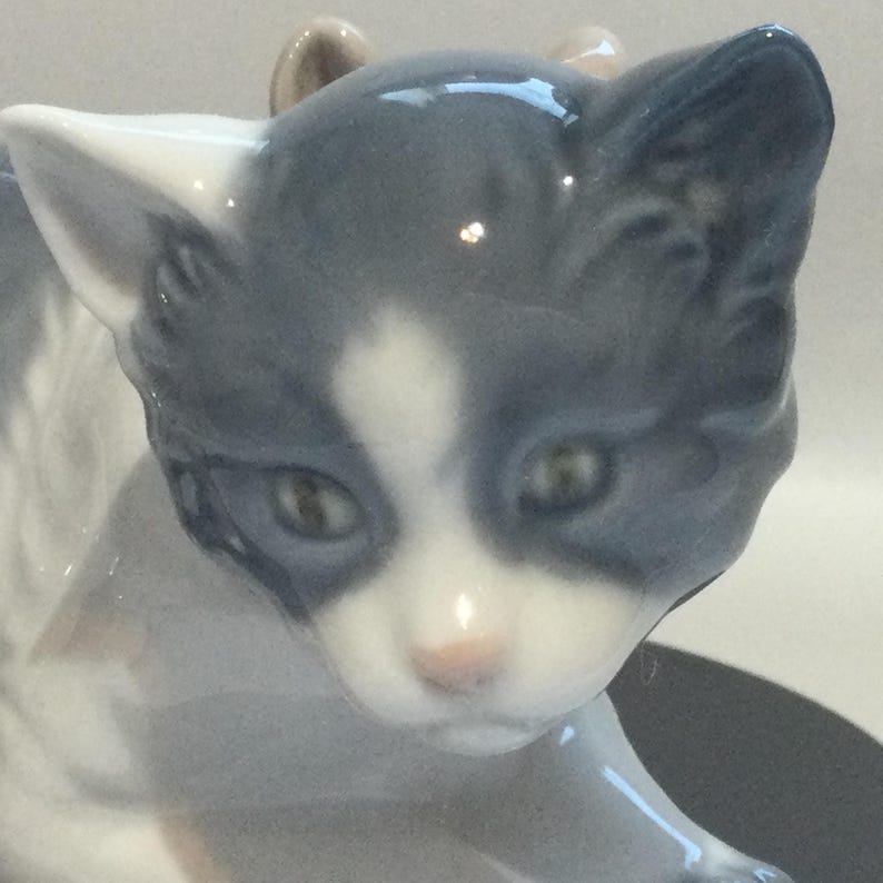 K&ouml;nnte beinhalten: Eine Keramikfigur einer Katze mit grau-wei&szlig;em Fell. Die Katze hat einen wei&szlig;en Fleck auf der Stirn und eine rosa Nase. Die Figur ist detailliert mit realistischen Augen und Ohren und ist ein dekorativer Artikel.