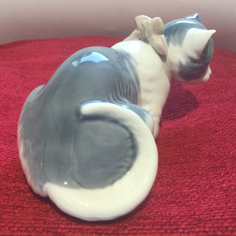 K&ouml;nnte beinhalten: Eine Keramik-Katzenfigur in sitzender Position, mit grau-wei&szlig;em Farbschema. Die Katze hat eine Schleife auf dem R&uuml;cken und einen eingerollten Schwanz. Die Figur steht auf einer roten Oberfl&auml;che.