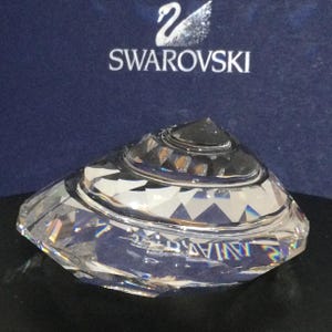Op de afbeelding: Een heldere kristallen schelp sculptuur met een draaiend ontwerp. Het beeld is gemaakt door Swarovski en wordt getoond op een blauwe achtergrond.