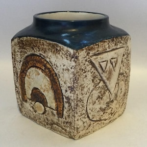 May include: A square ceramic vase with a dark blue glazed rim and a textured, off-white body. One side features a raised brown arch design with a circular base, while the adjacent side displays a raised triangle containing two smaller triangles.