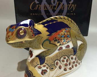 Puede incluir: Una figurita de camaleón de cerámica dorada, verde, azul y roja con un diseño detallado. El camaleón está posado sobre una roca con un fondo azul y dorado. La figurita está hecha por Royal Crown Derby, English Fine Bone China.