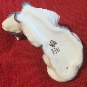 K&ouml;nnte beinhalten: Eine wei&szlig;e Keramikfigur einer Katze mit grauen Akzenten. Die Unterseite der Figur weist ein Logo und Text auf. Die Katze ist in einer gekr&uuml;mmten Position, mit Schwanz und Kopf sichtbar. Auf rotem Untergrund.