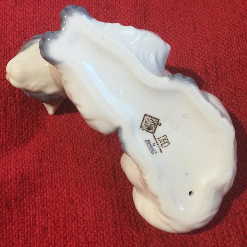 K&ouml;nnte beinhalten: Eine wei&szlig;e Keramikfigur einer Katze mit grauen Akzenten. Die Unterseite der Figur weist ein Logo und Text auf. Die Katze ist in einer gekr&uuml;mmten Position, mit Schwanz und Kopf sichtbar. Auf rotem Untergrund.