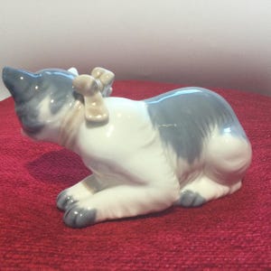 K&ouml;nnte beinhalten: Eine Keramik-Katzenfigur in Ruheposition, mit grau-wei&szlig;em Farbschema und einer kleinen Schleife. Die Ohren und Pfoten der Katze sind grau, w&auml;hrend ihr K&ouml;rper &uuml;berwiegend wei&szlig; ist. Die Figur ist ein dekoratives Objekt.