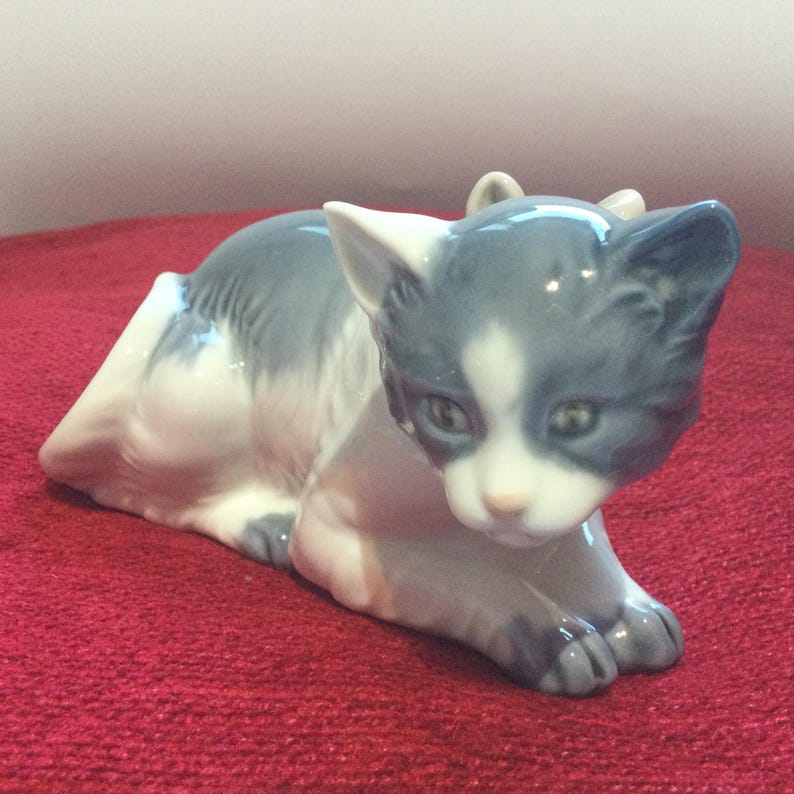 K&ouml;nnte beinhalten: Porzellanfigur einer ruhenden Katze. Die Katze hat ein grau-wei&szlig;es Fell mit detaillierten Gesichtsz&uuml;gen. Die Figur steht vor einem roten Hintergrund.