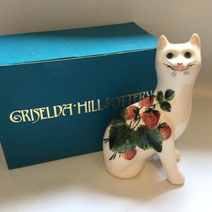 以下が含まれることがあります： 赤いイチゴと緑の葉の模様が描かれた白い陶器製の猫の置物。その横には、前面に金色の文字で「GRISELDA HILL POTTERY」と印刷された青緑色の長方形の箱が置かれています。
