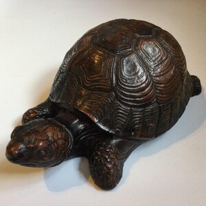 May include: A detailed, brown tortoise sculpture with a textured shell and detailed head and limbs. The sculpture is made of a dark brown material and sits on a white surface. The tortoise is a decorative item.