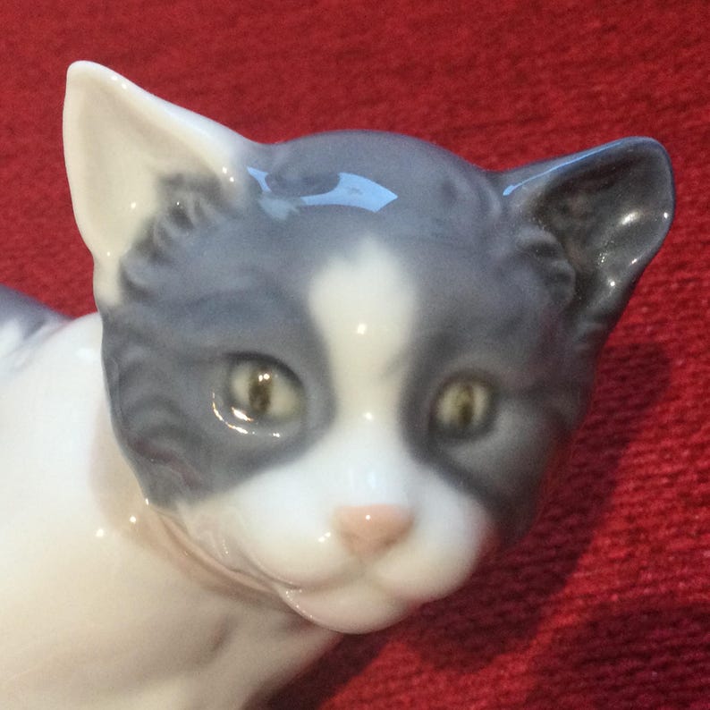 K&ouml;nnte beinhalten: Eine Keramikfigur einer Katze mit grau-wei&szlig;em Fell. Das Gesicht der Katze ist mit gr&uuml;nen Augen und einer rosa Nase detailliert. Die Figur befindet sich vor einem roten Hintergrund.