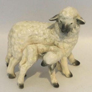 May include: A ceramic figurine depicting a mother sheep standing with a small lamb. The sheep have a glossy cream-coloured finish with textured wool details, dark grey ears, faces, and hooves, capturing a realistic pose of the two animals together.