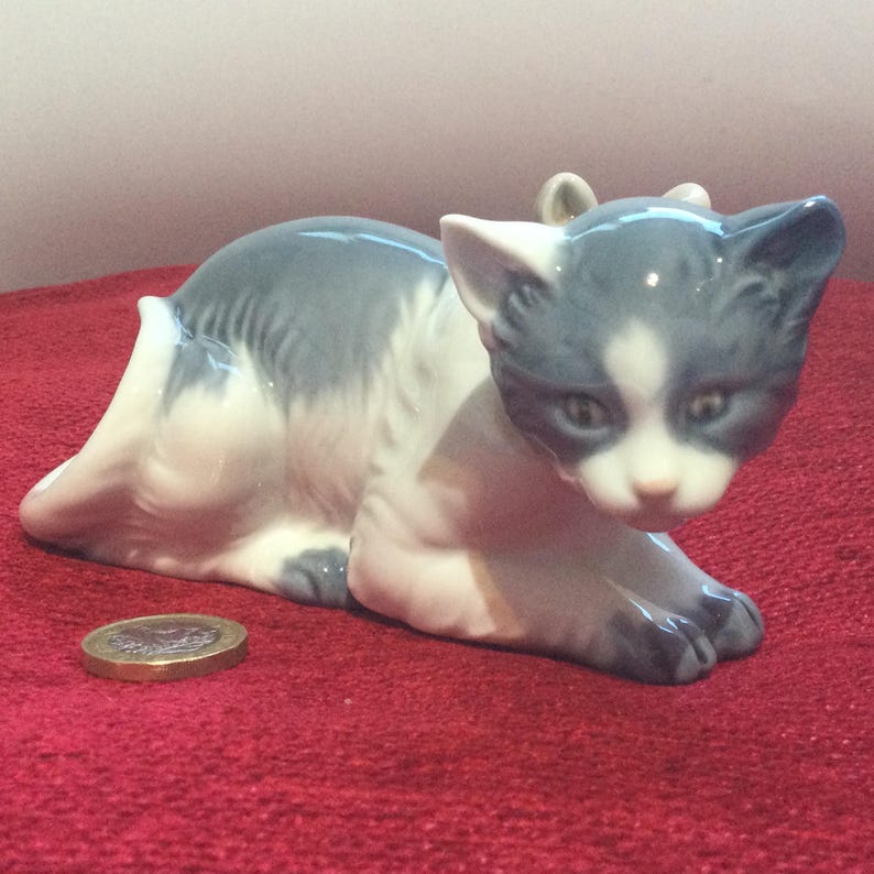 K&ouml;nnte beinhalten: Eine Keramikfigur einer Katze in Ruheposition. Die Katze ist haupts&auml;chlich wei&szlig; mit grauen Markierungen auf Kopf, R&uuml;cken und Pfoten. Die Figur steht auf einer roten Oberfl&auml;che. Eine M&uuml;nze liegt neben der Katze.