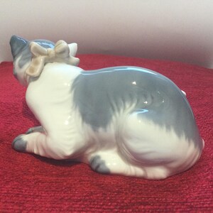K&ouml;nnte beinhalten: Eine Keramikfigur einer Katze in Ruheposition. Die Katze hat ein grau-wei&szlig;es Fell und eine kleine Schleife auf dem Kopf. Die Figur steht auf einer roten Oberfl&auml;che.