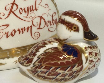 Puede incluir: Figura de pato de porcelana con cabeza y alas marrones, detalles dorados y azules. El pato está sobre un fondo blanco, con una vista parcial de una pieza de cerámica blanca con las palabras "Royal Crown Derby" en escritura roja.