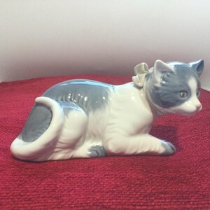 K&ouml;nnte beinhalten: Eine Keramikfigur einer Katze in Ruheposition. Die Katze ist &uuml;berwiegend wei&szlig; mit grauen Markierungen an Kopf, R&uuml;cken und Schwanz. Ein kleines Schleifchen ziert ihren Hals. Die Figur steht auf einer roten Oberfl&auml;che.