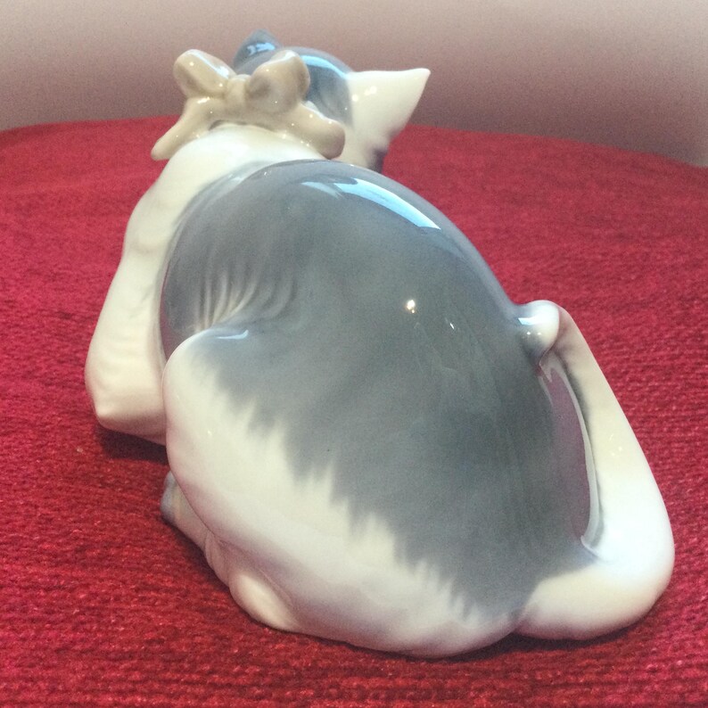 K&ouml;nnte beinhalten: Keramikfigur einer sitzenden Katze, von hinten gesehen. Die Katze hat ein grau-wei&szlig;es Fell und eine Schleife auf dem Kopf. Die Figur steht auf einer roten Oberfl&auml;che.
