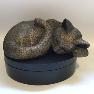 Può includere: Una scultura color bronzo di un gatto raggomitolato in posizione di riposo con dettagli del pelo testurizzati. La figura del gatto poggia su una base nera opaca, liscia e circolare, caratterizzata da una distinta cucitura orizzontale.