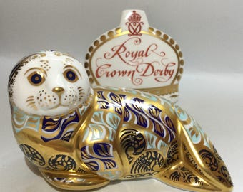 Puede incluir: Una figurita de foca Royal Crown Derby con detalles en azul, dorado y blanco. La foca está tumbada boca abajo con la cabeza girada hacia un lado. La figurita está hecha de porcelana y está en perfecto estado.