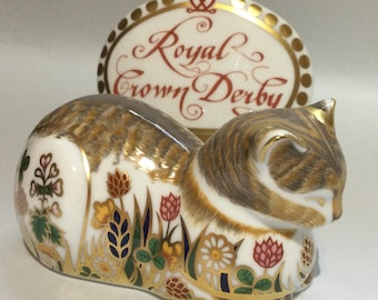 Puede incluir: Una figura de gato de porcelana blanca con rayas atigradas marrones y patrones florales coloridos en rojo, amarillo y verde. Detrás del gato hay una placa de porcelana blanca con borde dorado y el texto rojo Royal Crown Derby.