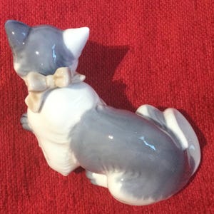 K&ouml;nnte beinhalten: Eine Keramikfigur einer Katze, haupts&auml;chlich grau und wei&szlig;, mit einer beigen Schleife. Die Katze sitzt, mit dem R&uuml;cken zum Betrachter. Die Figur hat eine gl&auml;nzende Oberfl&auml;che.