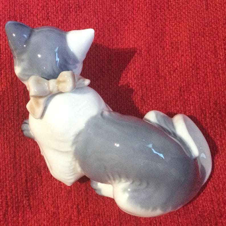 K&ouml;nnte beinhalten: Eine Keramikfigur einer Katze, haupts&auml;chlich grau und wei&szlig;, mit einer beigen Schleife. Die Katze sitzt, mit dem R&uuml;cken zum Betrachter. Die Figur hat eine gl&auml;nzende Oberfl&auml;che.