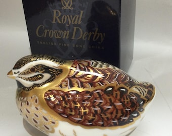 Puede incluir: Una figurita de porcelana de una codorniz marrón y blanca con detalles dorados. La figurita está sentada sobre una superficie blanca y delante de una caja azul con las palabras "Royal Crown Derby English Fine Bone China" impresas en ella.