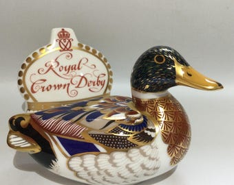 Puede incluir: Figura de pato de porcelana Royal Crown Derby con intrincados detalles dorados, azules y marrones. El pato tiene una cabeza verde y negra, un pico amarillo y plumas blancas. Un letrero de Royal Crown Derby está detrás del pato.