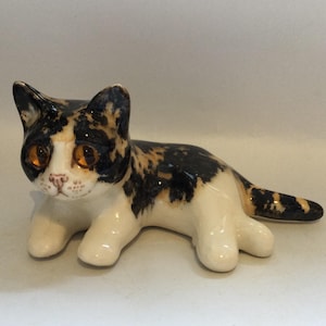 以下が含まれることがあります： 黒、オレンジ、白の斑点模様の三毛猫の陶器製フィギュア。横たわった姿勢で、大きな光沢のある琥珀色の目、小さなピンクの鼻、白い口元に描かれたひげが特徴です。