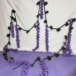 May include: Crocheted wisteria garland in shades of purple and green. The garland features hanging clusters of purple flowers and green leaves, creating a decorative floral design. The handmade garland is displayed against a white and purple background.