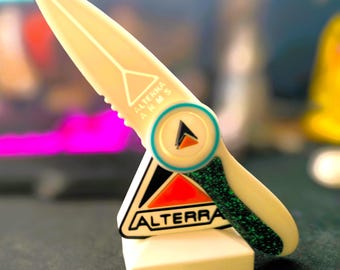 Könnte beinhalten: Ein weißes Plastikmesser mit einem grünen Glitzergriff und einem schwarzen und roten Dreieckslogo mit der Aufschrift "ALTERRA". Das Messer hat einen weißen Kreis mit einem blauen Rand und einem schwarzen Dreieck darin. Der Text auf dem Messer lautet "ALTERRA ARM S".