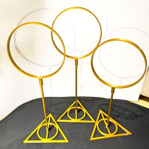 May include: Three gold-colored decorative stands. Each stand features a circular hoop atop a slender pole, connected to a triangular base with a circular design. The stands are arranged on a dark surface, creating a visually appealing display.