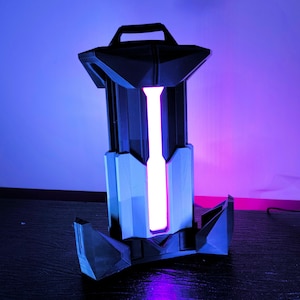 Puede incluir: Una lámpara negra y gris impresa en 3D con una luz rosa en su interior. La lámpara tiene un asa en la parte superior y una base con un diseño geométrico.